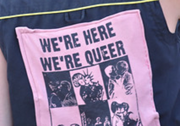 Close-up of a person wearing a sleeveless black shirt with a pink patch on the back that reads "WE'RE HERE WE'RE QUEER WE RIOT!" in bold text.