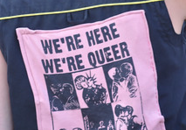 Back of a person's shirt with a large patch reading, "We're here. We're Queer. We Riot!"