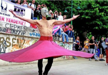 A shirtless person wearing a gas mask spins in a flowing pink skirt during a public protest, with onlookers and banners in the background.