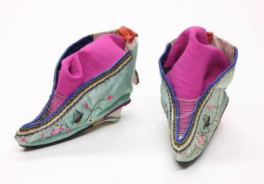 A pair of traditional Chinese lotus shoes with teal silk, floral embroidery, and upturned pointed toes. The small shoes for bound feet are lined with vibrant pink fabric, contrasting with blue trim.