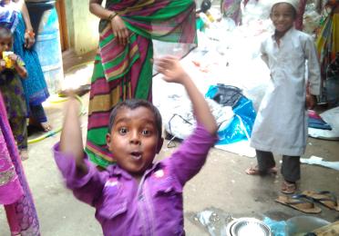 A smiling woman in a saree stands in an alleyway, with a boy in a purple shirt jumping energetically in the foreground.