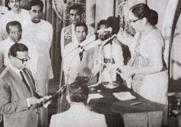 A black-and-white photograph of Sri Lanka’s 1972 ceremony declaring its first republican constitution. A woman administers an oath at a podium as officials stand nearby, marking the historic moment.