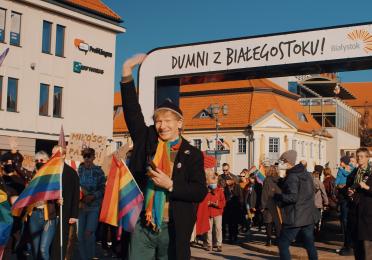 A person waves while leading a group in a pride parade under an arch with "Dumni z Białegostoku!" People are holding rainbow flags in a sunlit urban setting.