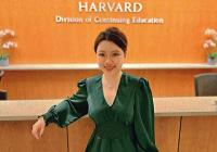 Upper body shot of Alva He wearing a green blouse, khaki pants, and her hair in a bun. She is standing in front of a desk with the emblem for Harvard Division of Continuing Education behind her.