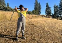 Keila Taylor wearing a yellow shirt and overalls standing in a field with her legs spread and firmly planted and her hands clasped behind her head. She's looking up at the sky. 
