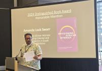 A person stands at a podium speaking, with a presentation slide behind them about the 2024 Distinguished Book Award Honorable Mention for "Envisioning African Intersex" by Amanda Lock Swarr.