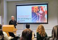 Shelley Pryde stands at a podium presenting during Scholars' Studio. The presentation slide behind them features the title, 'The art of drag in Shanghai,' along with the quote, 'We’re all born naked and the rest is Communism.' A vibrant image of a drag performer is displayed on the slide.