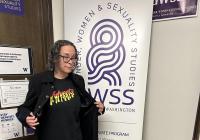 Maria Breaux standing beside a GWSS banner, opening her jacket to show a ‘Vulveeta’ T-shirt.