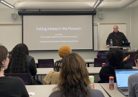 Eric Villiers standing behind a podium in front of a classroom of students. On the screen behind him, the title of his presentation, "Falling Asleep in the Museum"