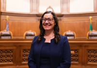 Colleen Melody smiles while seated in a wood-paneled courtroom, wearing glasses and a navy suit. Judicial chairs and the U.S. and Washington State flags appear in the background.