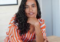 Portrait of artist Chloë Bass smiling at the camera, wearing an orange and gray striped shirt against a neutral background.