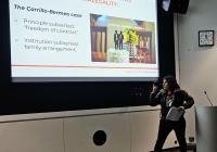 Eli stands in front of a projector screen pointing to the image displayed and presenting