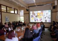 A group of students and faculty sit around a table and in chairs along the wall looking toward a projector screen as a presentation is being shown