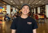 Upper body shot of Kai Mai in a black tshirt with a baseball cap on his head. Kai is a young Asian male with light skin and is smiling at the camera.