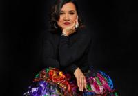 Martha Gonzalez seated against a dark background, wearing a vibrant multicolored skirt with embroidered floral patterns and a black long-sleeved top, resting her chin on her hand with a thoughtful expression.