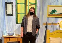 A man standing in the center of a stage designed to look like a bedroom with a chair, desk, bed, and artwork on the walls. The man with shoulder-length brown hair is wearing a sweater over a button-up shirt, black pants and shoes, and a mask on his face.