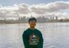 Full body image of Kenji Fujinaka sitting on a cement wall with the Seattle skyline in the background, wearing a green sweatshirt, black running pants, and white gym shoes and smiling at the camera 