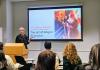 Shelley Pryde stands at a podium presenting during Scholars' Studio. The presentation slide behind them features the title, 'The art of drag in Shanghai,' along with the quote, 'We’re all born naked and the rest is Communism.' A vibrant image of a drag performer is displayed on the slide.