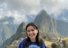  Fiona Rivera, an undergraduate student, stands at Machu Picchu, wearing a blue and green tie-dye Earth shirt. The stone terraces and misty mountains are visible behind her.