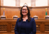 Colleen Melody smiles while seated in a wood-paneled courtroom, wearing glasses and a navy suit. Judicial chairs and the U.S. and Washington State flags appear in the background.