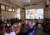 A group of students and faculty sit around a table and in chairs along the wall looking toward a projector screen as a presentation is being shown