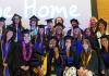 Students of the Class of 2026 stands and crouches for a group photo wearing their black graduation cap and gown in front of a projector screen