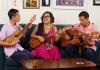 Image of woman (Martha Gonzalez) sitting in the middle of a couch playing a guitar with her son and husband on either side of her also playing guitars. 
