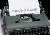 A typewriter on a plain desk with a sheet of paper sticking out. The paper reads "Funding Round" in large print, all caps, across the center of the page.