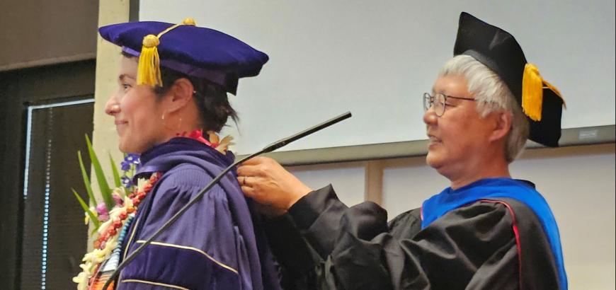 Profile of two people in graduation caps and gowns. One is behind the other adjusting the doctoral hood that has just been placed over the other's head.