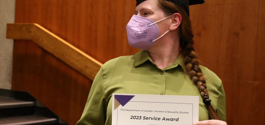 Upper body shot of Ameli Cyr. She is wearing a graduation cap, her hair in a long brain, and a face mask. She is looking to her left while holding up the certificate for 2023 GWSS Service Award.