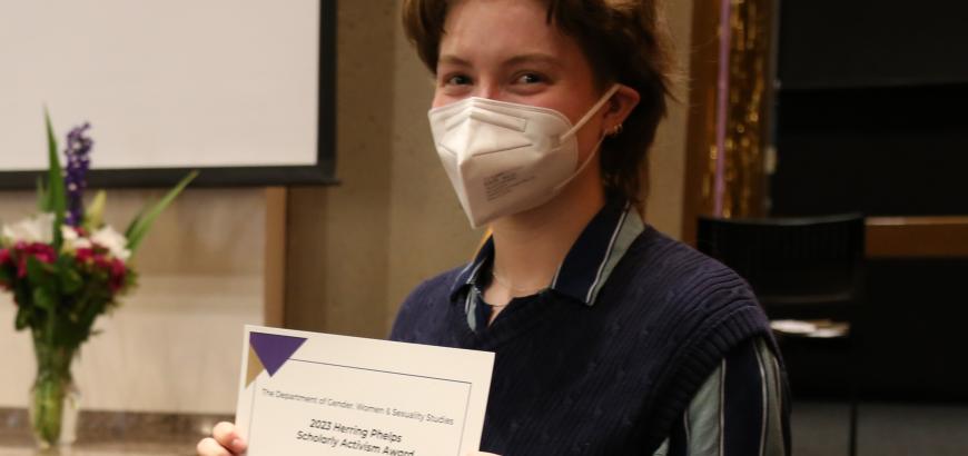 Upper body shot of Daisy Federspiel-Baier. She has short brown hair, is wearing a face mask and smiling at the camera while holding up a copy of her award certificate.