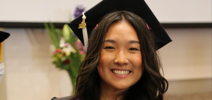 Upper body shot of Deborah Jin. She has on full graduation regalia and is holding up the certificate for her B.A. in Gender, Women &amp; Sexuality Studies.