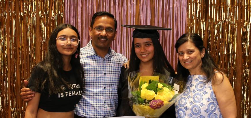 Photobooth image of Deeya Sharma with sister and parents. She is holding flowers and her graduation certificate. The banner above her head reads &quot;congrats&quot; with a gold tinsel backdrop.