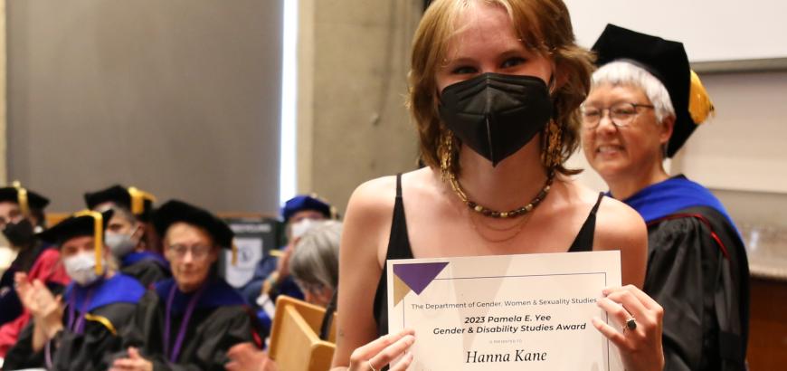 Upper body shot of Hanna Kane smiling at camera and holding award certificate. Background left: seated faculty members. Background right: Professor Shirley Yee standing and smiling towards Hanna.