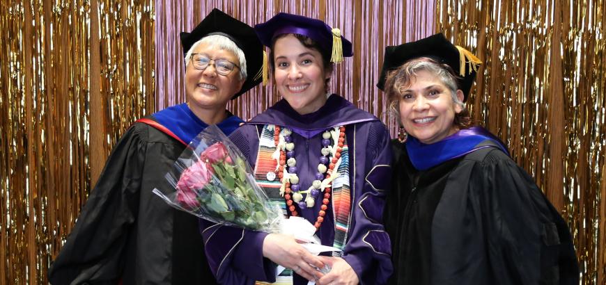Three women in graduation caps and gowns smiling at the camera and standing in front of purple and gold tinsel with a banner reading &quot;congrats&quot; overhead.