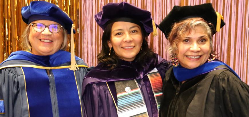 Upper body image of three women in graduation caps and gown smiling for the camera. The background is pink and gold tinsel.