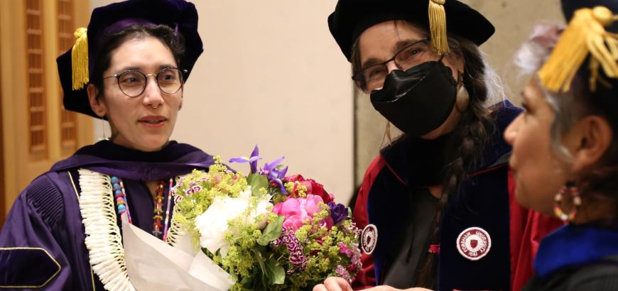 Group of three people in graduation caps and gowns celebrating. The person on the left is holding a bouquet of flowers, the one in the middle is smiling at the camera, and the one on the right is snacking from a plate of fruit.