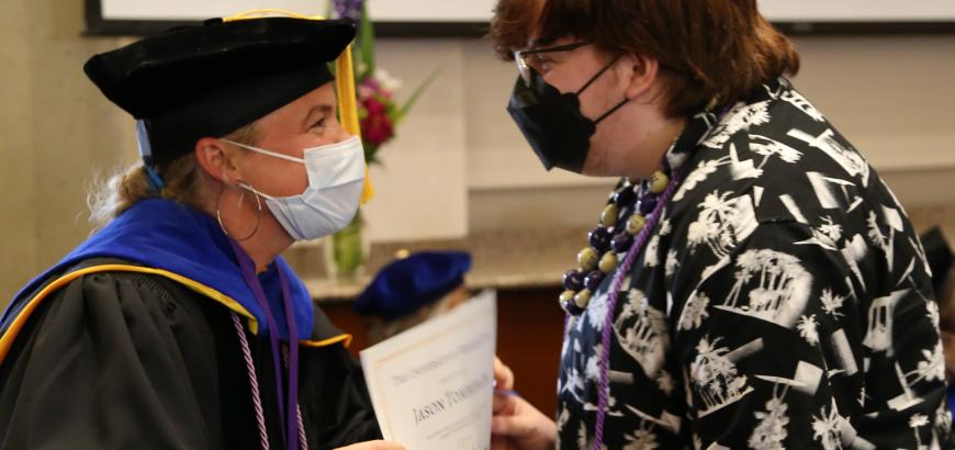 Side view of a professor in graduation cap and gown handing a certificate to a graduating student.