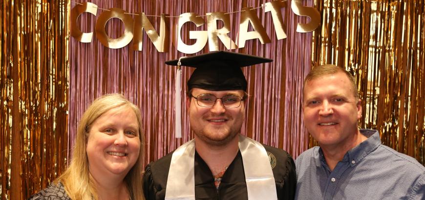 Parents standing on either side of their graduating son. The son is wearing graduation cap and gown. Background gold and pink tinsel with banner reading &quot;congrats&quot; hanging above their heads.
