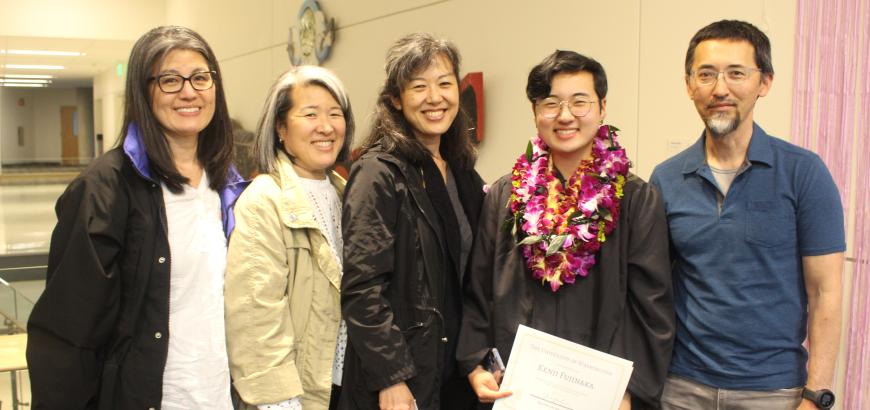 A group of 5 people in a row who appear to be related. The second from the right is wearing a graduation robe, banner and a lei.