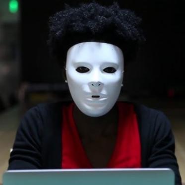 A person wearing a white expressionless mask sits at a laptop in a dimly lit room. They have short curly hair and are dressed in a red shirt and dark cardigan, creating a mysterious, unsettling atmosphere.