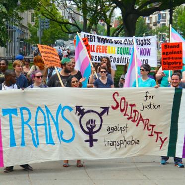 A group of people marching in a trans rights rally, holding trans pride flags and a large banner that reads “Trans Solidarity for justice against transphobia.”