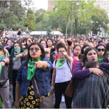 A large crowd of women, some with green bandanas and raised arms, participate in the feminist performance “A Rapist in Your Path” in Spain.