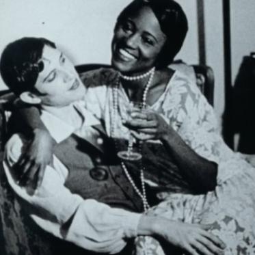 Black-and-white film still of two women seated closely together on a couch, smiling and holding drinks, with one woman’s arm wrapped around the other.