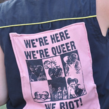 Close-up of a person wearing a sleeveless black shirt with a pink patch on the back that reads &quot;WE'RE HERE WE'RE QUEER WE RIOT!&quot; in bold text.