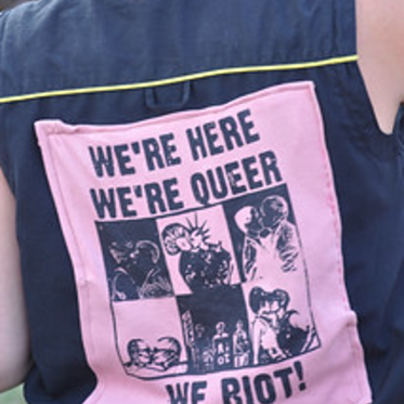 Back of a person's shirt with a large patch reading, "We're here. We're Queer. We Riot!"