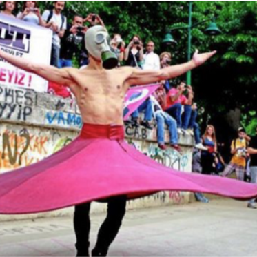 A shirtless person wearing a gas mask spins in a flowing pink skirt during a public protest, with onlookers and banners in the background.