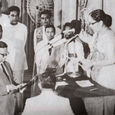 A black-and-white photograph of Sri Lanka’s 1972 ceremony declaring its first republican constitution. A woman administers an oath at a podium as officials stand nearby, marking the historic moment.