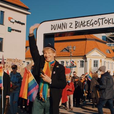 A person waves while leading a group in a pride parade under an arch with &quot;Dumni z Białegostoku!&quot; People are holding rainbow flags in a sunlit urban setting.